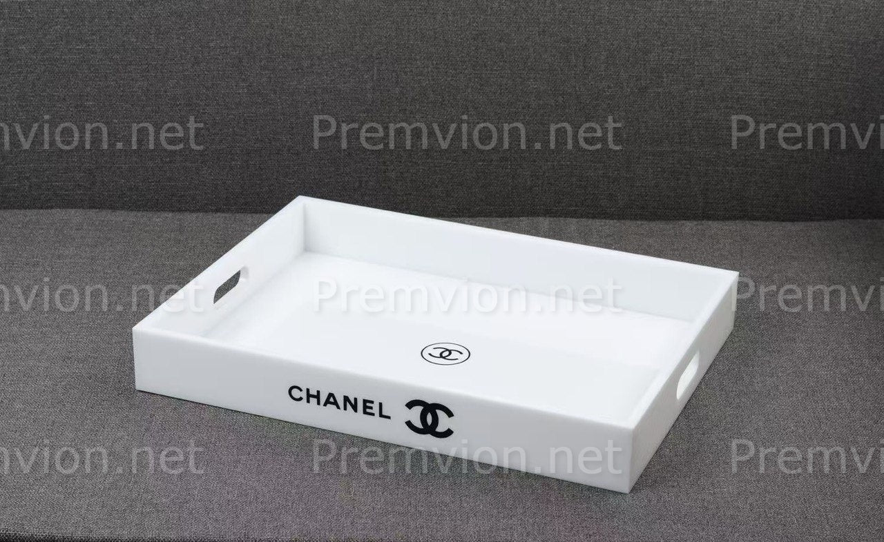 A white rectangular Chanel tray with side cutouts and a black printed Chanel logo on the front, shown on a dark gray textured background.