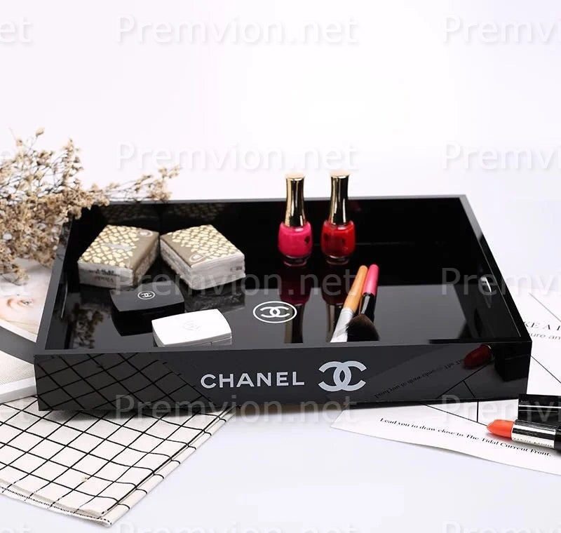 A black glossy Chanel cosmetic tray with the Chanel logo printed on the front, styled with nail polish bottles, lip gloss, and compact cases on a white surface.